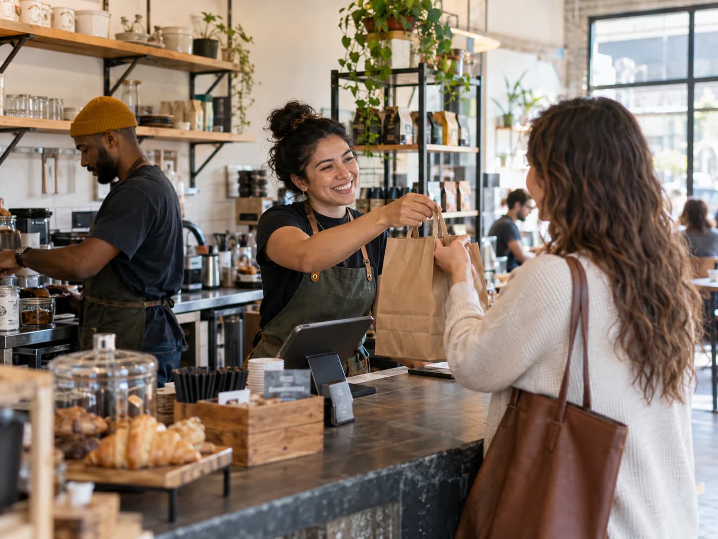 A neighborhood cafe employee handing an order to a customer while a teammate continues the workday nearby